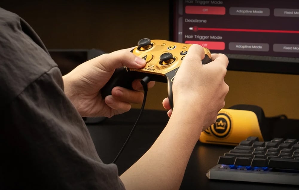 A person holding a wired game controller at a desk with a monitor and keyboard