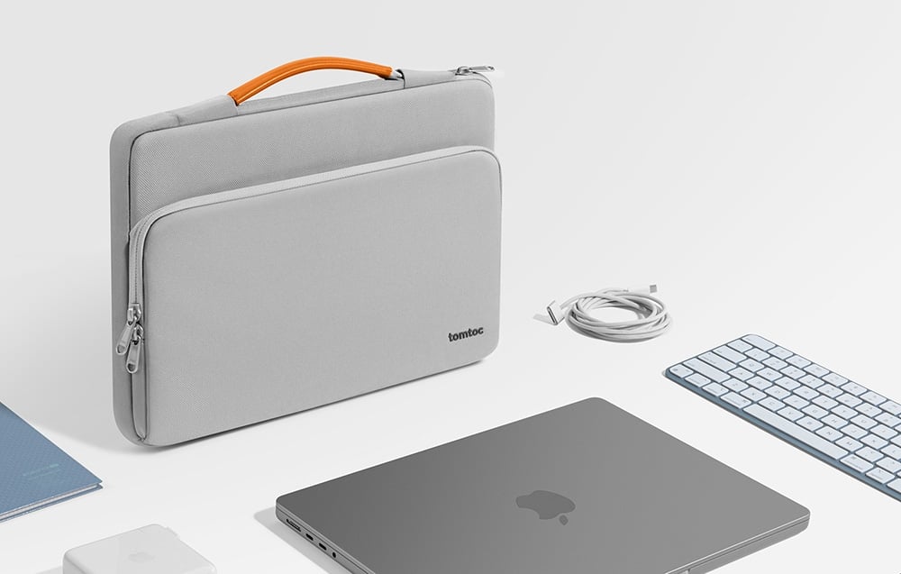 A light gray tomtoc bag stands on a white desk next to a laptop, keyboard, charger, cable, and notebook.