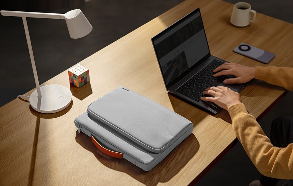 A top view of a desk where a man is typing on a laptop next to a tomtoc bag and a lit lamp.