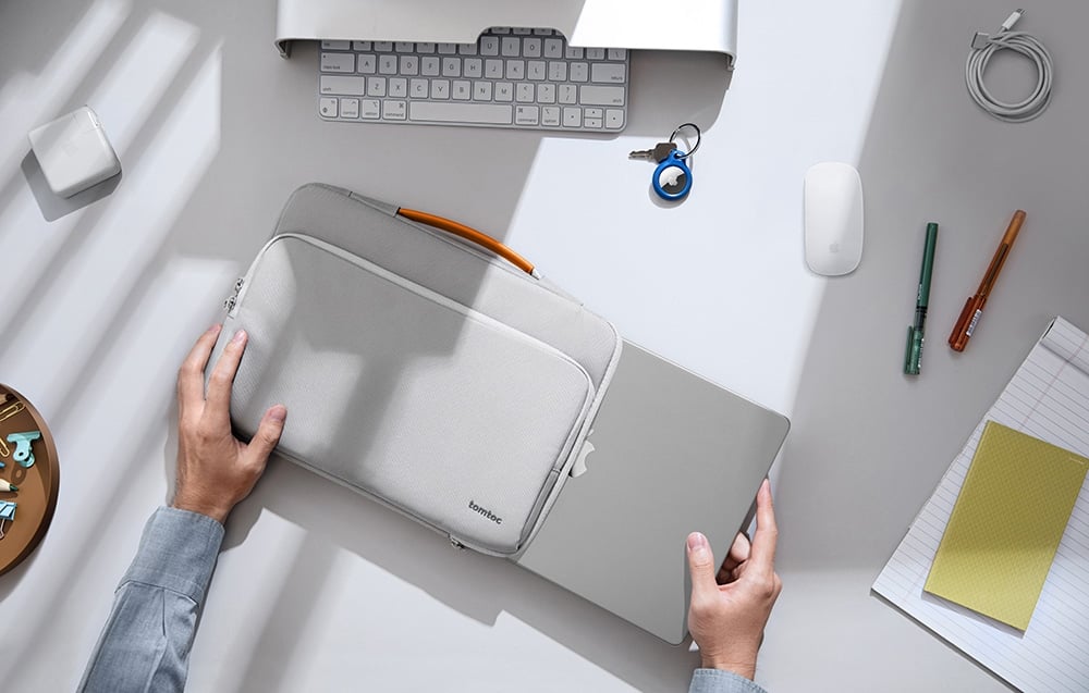 A woman sitting at a desk is putting a charger and cables into the front pocket of a light gray tomtoc bag.