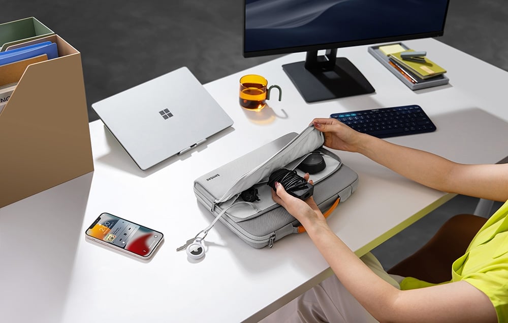 A woman sitting at a desk puts a charger and cables into the front pocket of the light gray tomtoc bag.