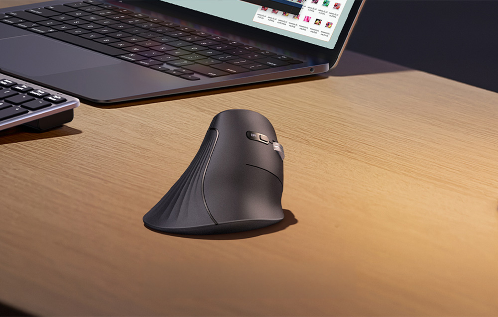 Black ergonomic vertical mouse standing on a wooden desk next to an open laptop