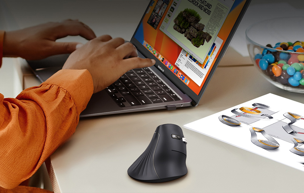 Black vertical mouse on a desk, with a person typing on a laptop next to a technical drawing