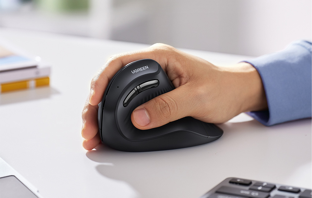 A hand holding a black ergonomic vertical mouse from UGREEN on a white desk