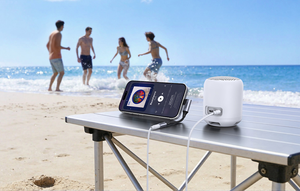 Telefon podłączony adapterem do głośnika na stole plażowym, z ludźmi bawiącymi się w tle