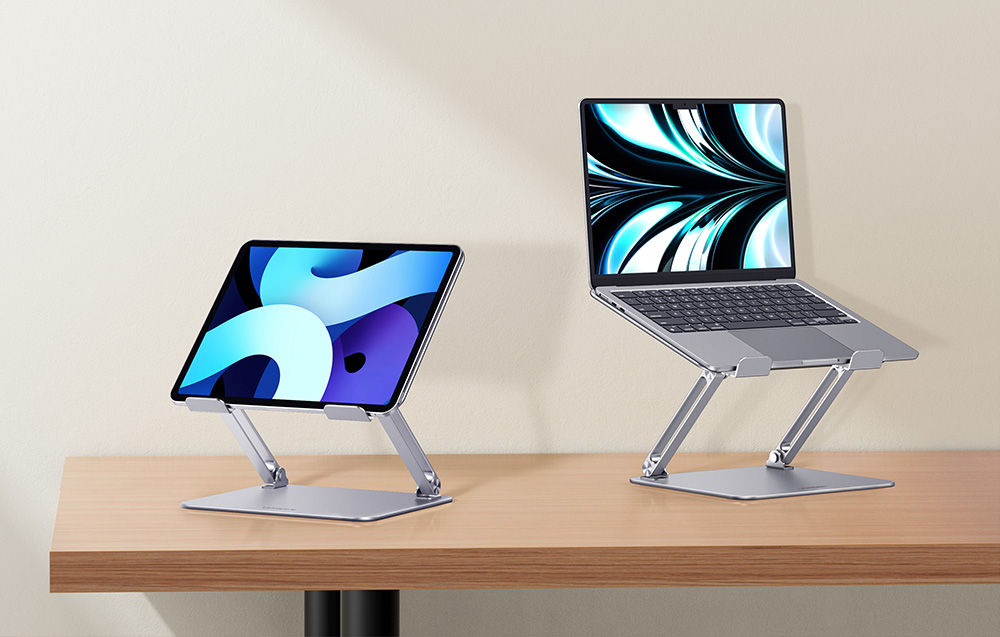 Tablet oraz laptop umieszczone na dwóch szarych, regulowanych podstawkach na biurku.