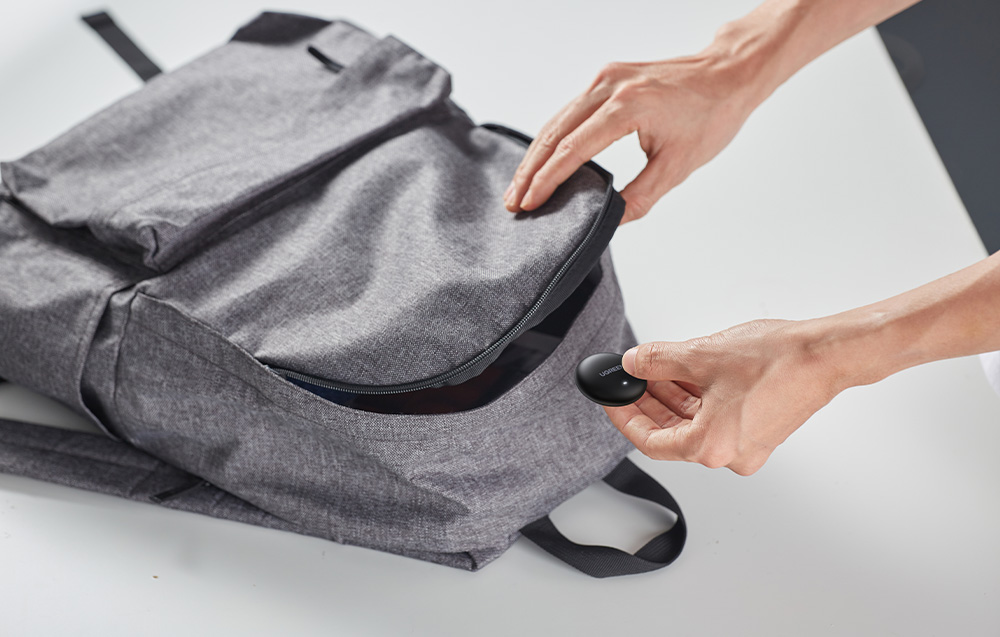 Person putting a black Ugreen locator into an open pocket of a gray backpack