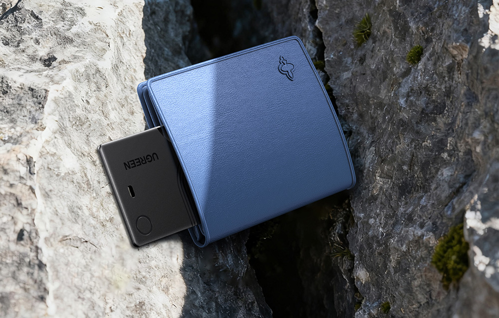 Black card-shaped Ugreen locator sticking out of a blue wallet lying on rocks