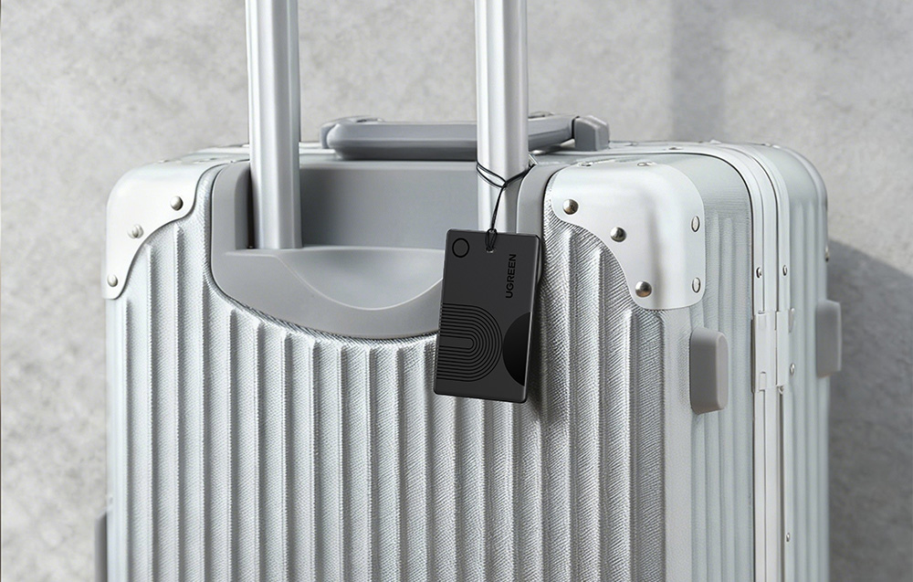 Black card-shaped Ugreen locator hanging from the handle of a silver suitcase