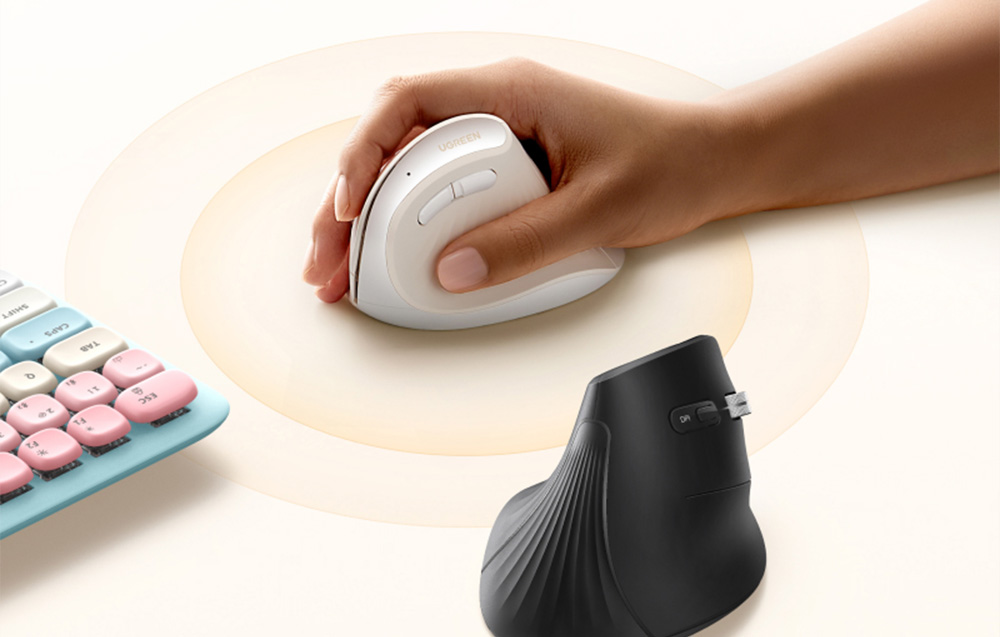 A hand holding a white vertical computer mouse next to a black model and a colorful keyboard