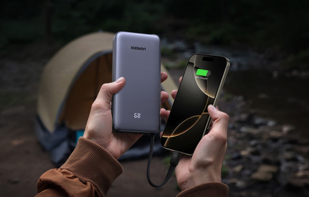 Two hands holding the gray Ugreen power bank and a smartphone connected with a cable against the backdrop of a camping tent