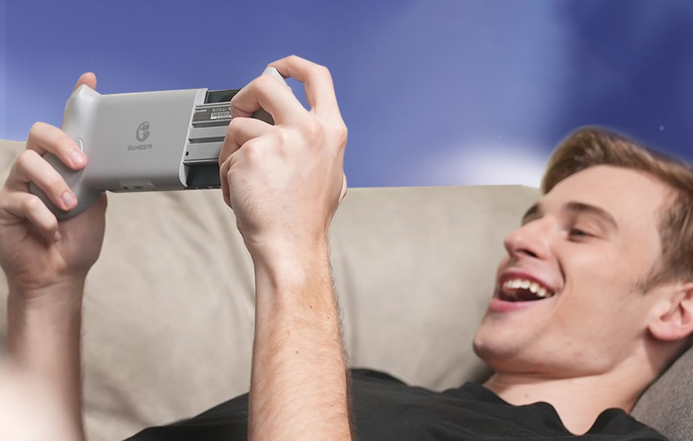 A smiling man lying on a couch playing a game with a controller connected to a smartphone