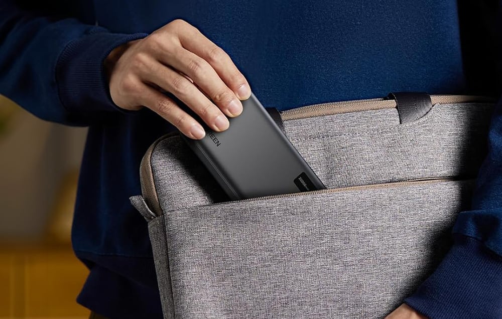 Close-up of a hand sliding the black power bank into the side pocket of a gray fabric laptop bag