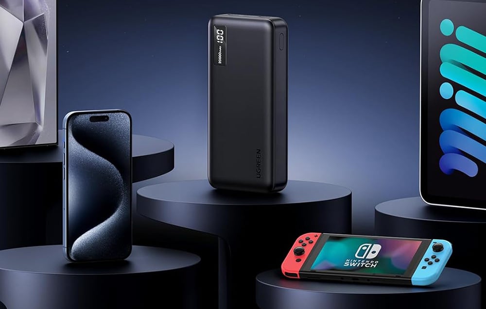 A black power bank with an activated display standing vertically on a round pedestal among other devices: smartphone, tablet, and gaming console with a colorful screen