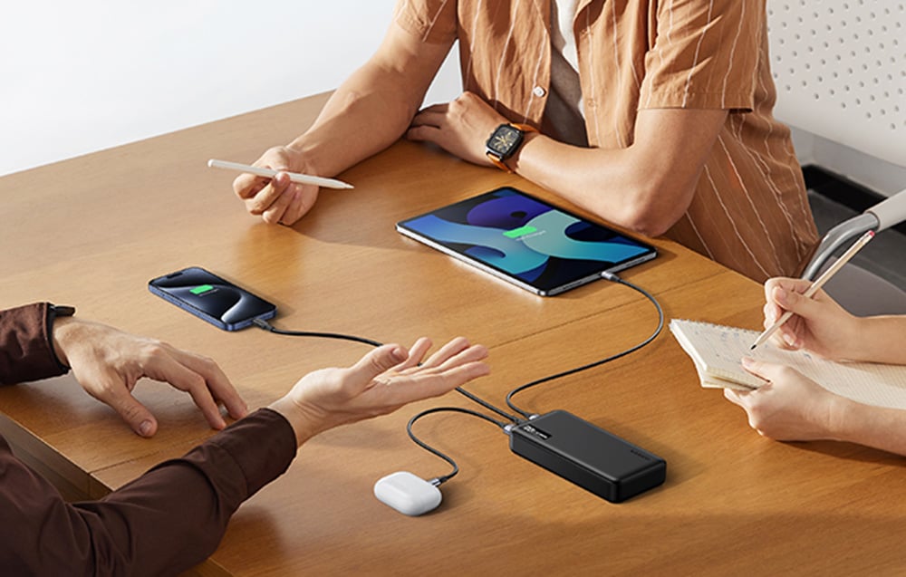 Several people sitting at a wooden table. A black power bank is on the countertop powering a tablet, smartphone, and wireless headphone case