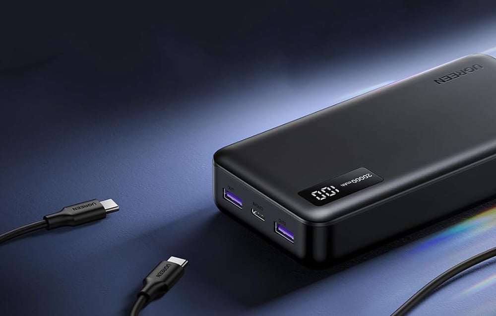 Black power bank with a digital display lying on a dark surface next to two detached USB-C cables